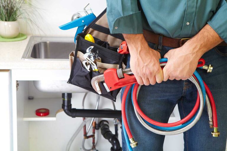 A plumber examining a wet, sunken patch of grass near a home’s foundation with plumbing tools nearby.