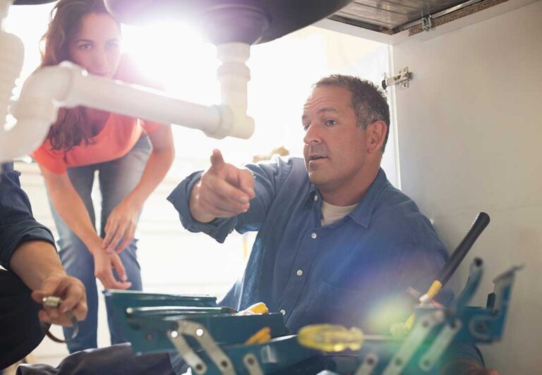 Homeowner checks a damp wall near a sink while holding a flashlight and examining exposed plumbing.