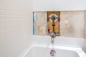 A homeowner checks a water meter while a plumber inspects a leaking toilet and damp bathroom cabinet.
